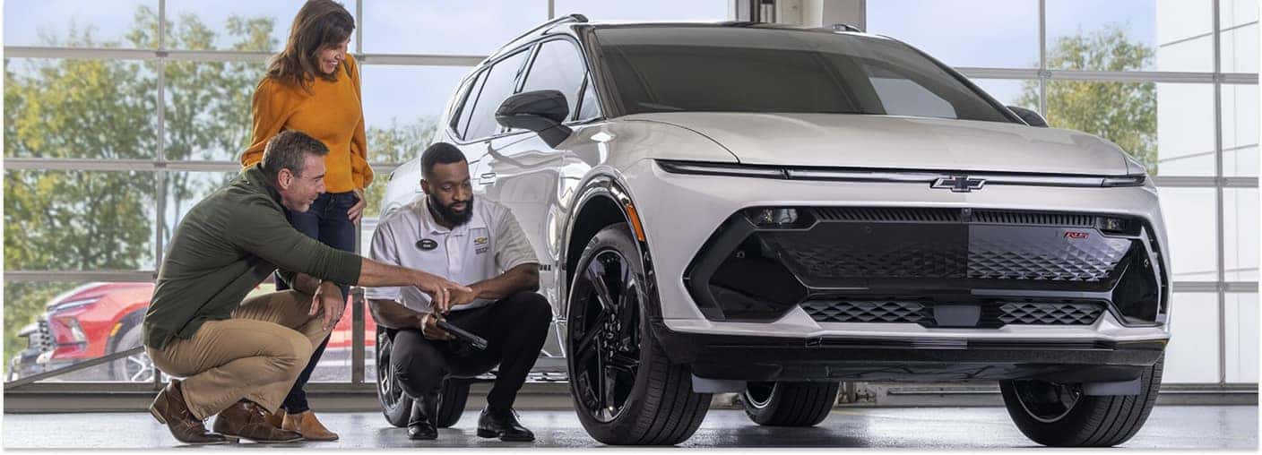 Technician inspecting the front tire of a white Chevrolet with customers in a service bay.