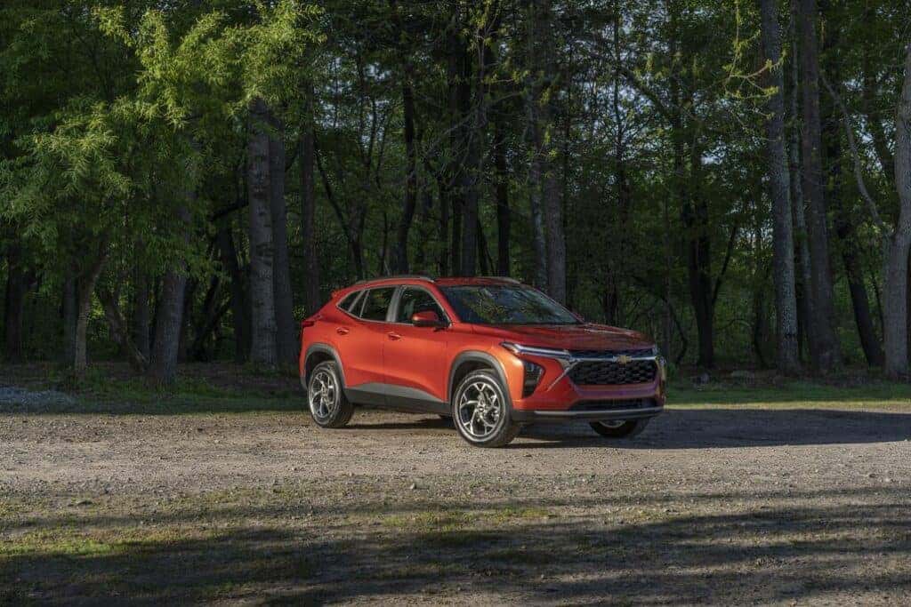 Front 3/4 view of 2024 Chevrolet Trax LT in Cayenne Orange Metallic parked on a gravel road in front of trees.