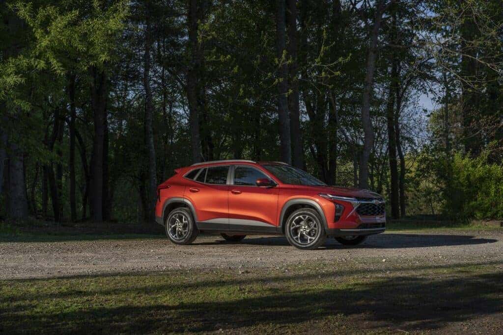Passenger’s side 7/8 view of 2024 Chevrolet Trax LT in Cayenne Orange Metallic parked on a gravel road in front of trees.