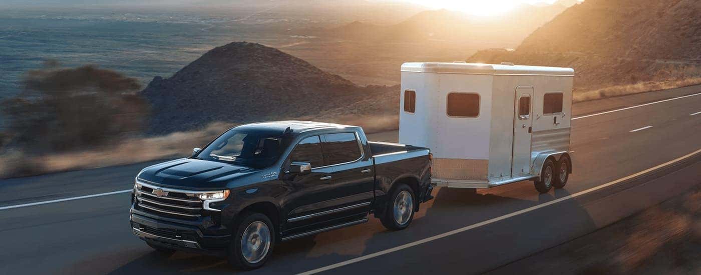 A grey 2023 Chevy Silverado 1500 High Country is shown towing a trailer on an open road.