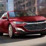 A red 2024 Chevy Malibu 2LT is driving on a city highway.