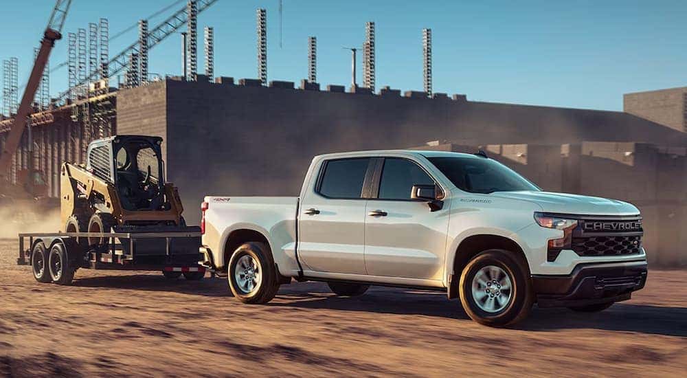 A white 2024 Chevy Silverado 1500 WT towing heavy equipment at a construction site.