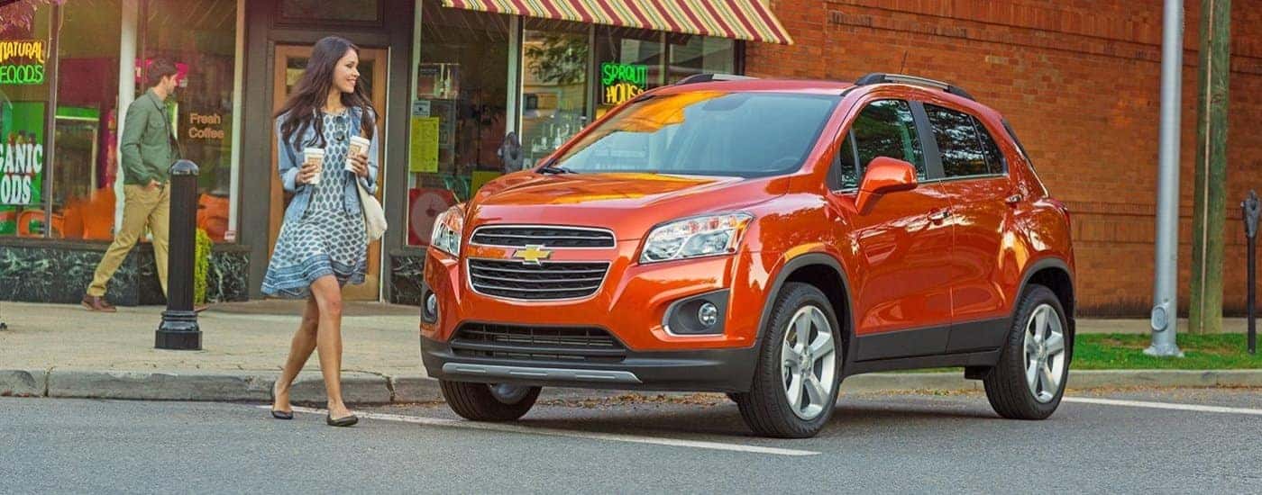 A person with coffee walking near an orange 2016 Chevy Trax.