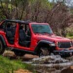 A red 2025 Jeep Wrangler Rubicon off-roading on a muddy path through a forest.
