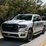 A silver 2025 Ram 1500 for sale is shown towing a trailer on a highway.