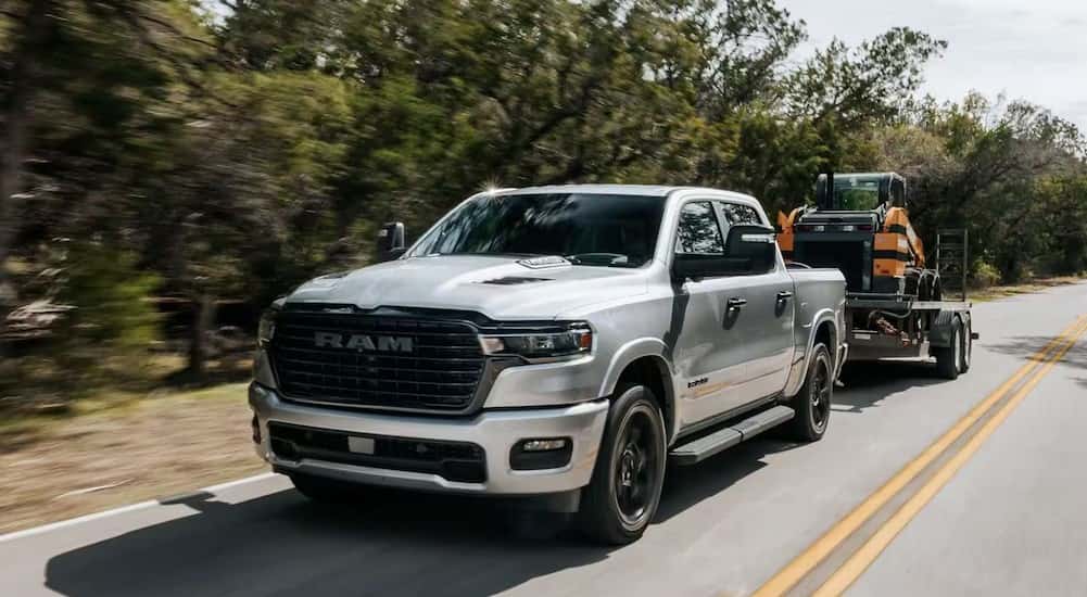 A silver 2025 Ram 1500 for sale is shown towing a trailer on a highway.