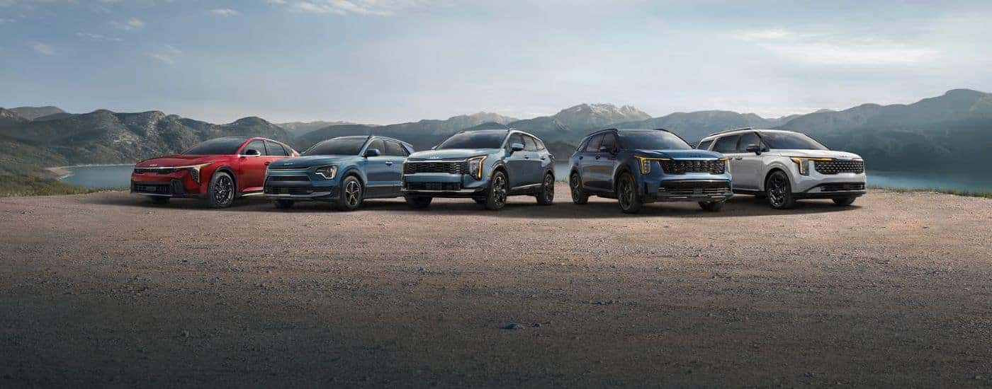 A line up of five 2026 Kia vehicles parked in front of a lake and mountains.