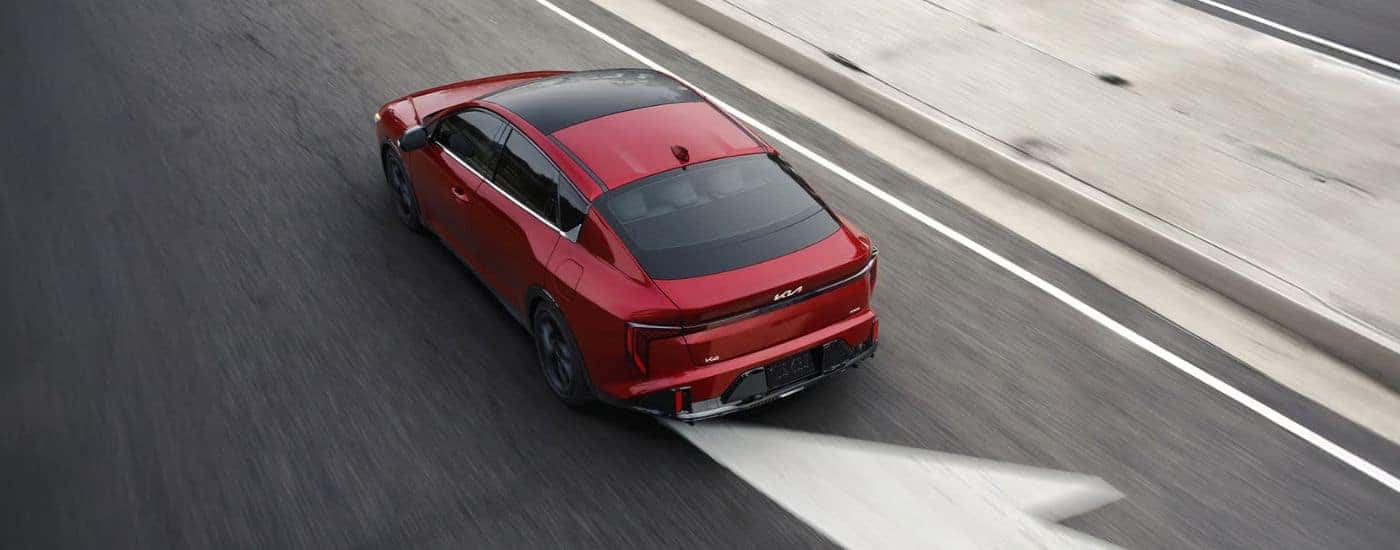 Rear view of a red 2026 Kia K4 GT Line Turbo with a wide sunroof driving on a paved road.