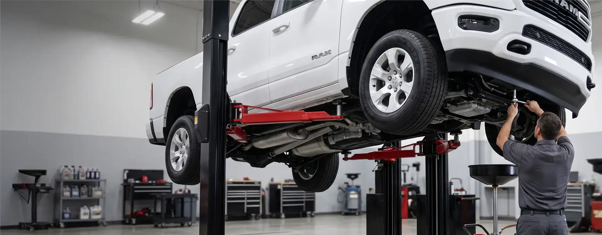 Technician working beneath a white RAM 1500 raised on a two-post lift, with a drain pan and waste oil caddy below in the Singing River CDJR service bay