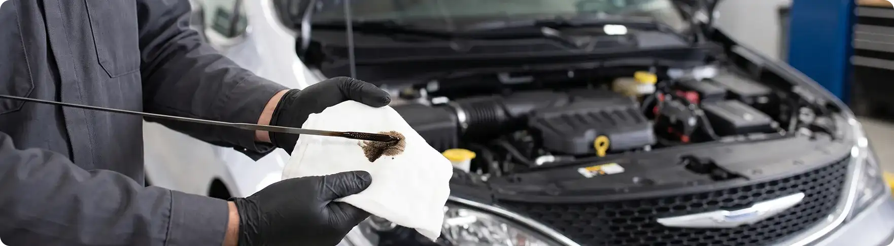 Gloved hand wiping a dipstick coated in dark used oil onto a white rag beside a silver Chrysler Pacifica with its hood open