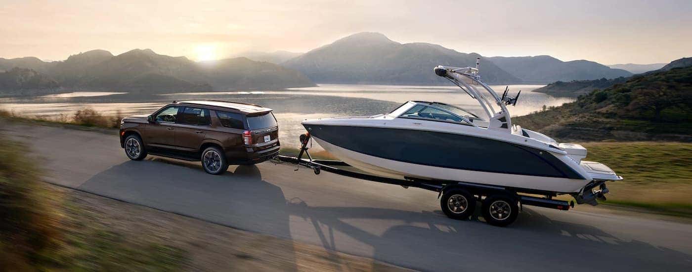 A brown 2022 Chevy Tahoe is shown towing a boar past a lake.