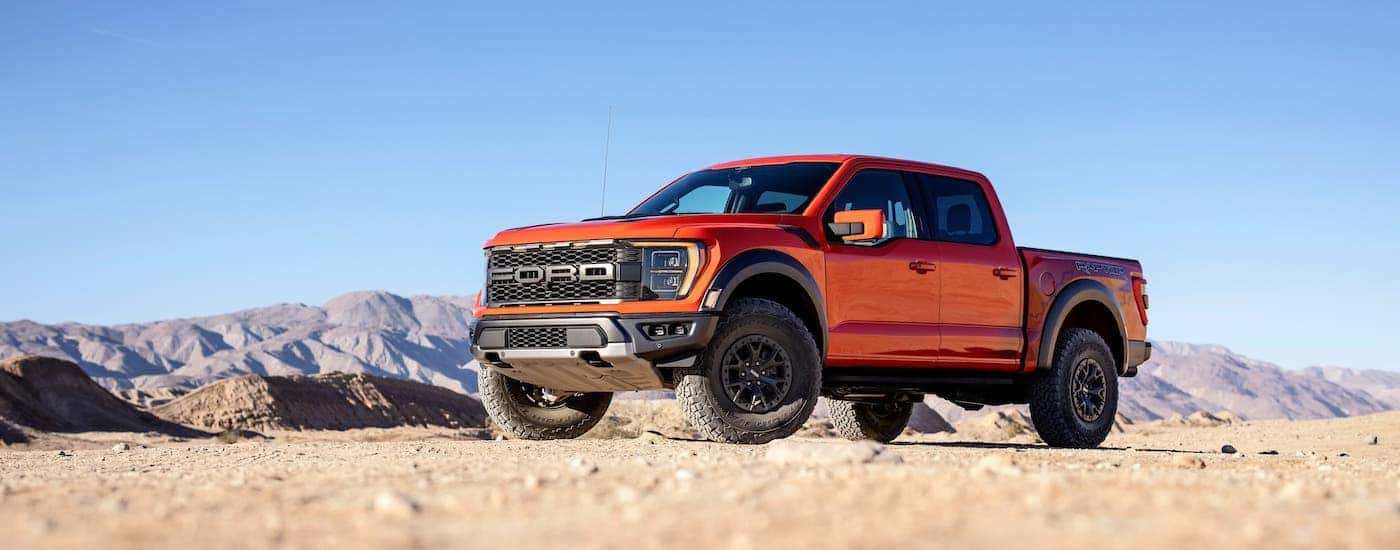 An orange 2021 Ford F-150 Raptor is shown parked in sand dunes.
