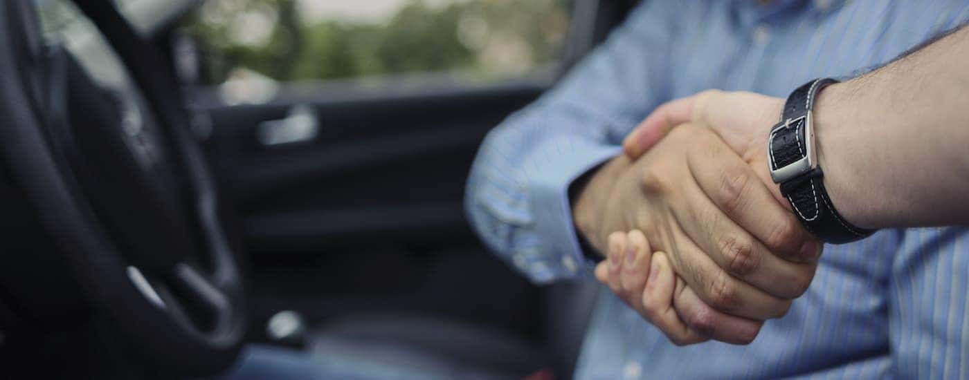 A person is shown shaking hands from the front seat of a vehicle.