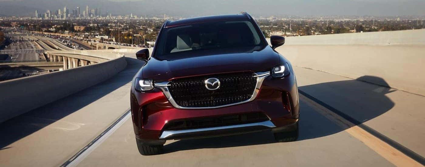 A maroon 2024 Mazda CX-90 PHEV is shown from the front driving on a highway.