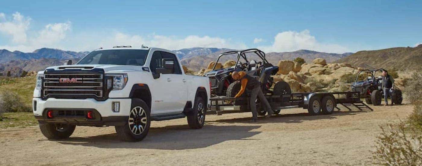A white 2020 GMC Sierra 2500 HD AT4 is shown from the front at an angle while unloading UTVs from a trailer.