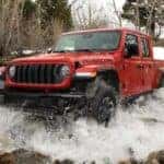 A red 2024 Jeep Gladiator Rubicon is shown driving over a shallow pond after visiting a dealer with trucks for sale.