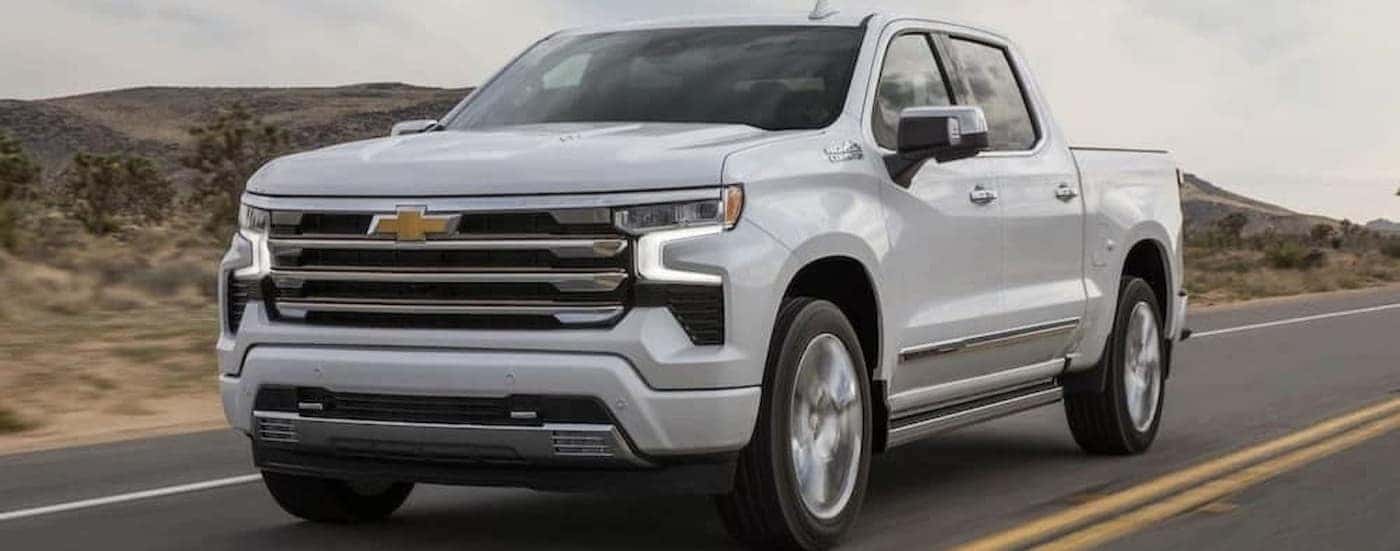 A white 2024 Chevy Silverado 1500 High Country is shown driving to view trucks for sale near Alamosa. 