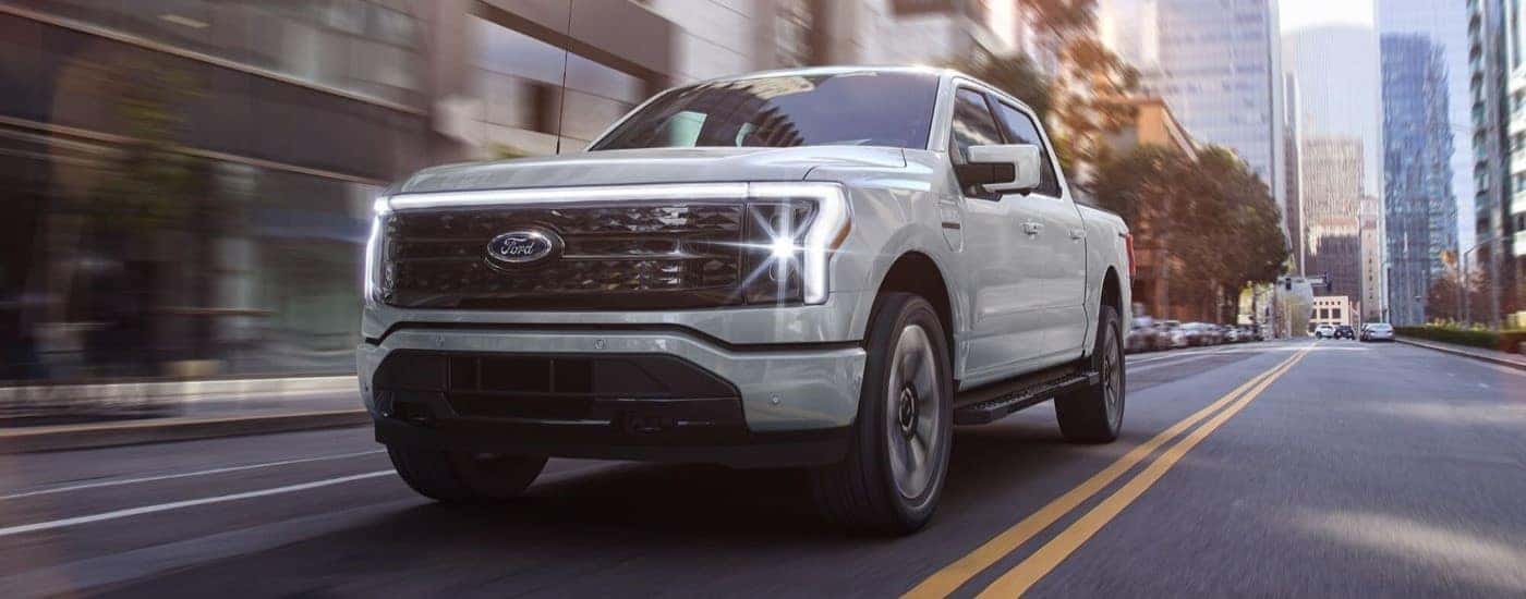 A white 2023 Ford F-150 Lightning is shown driving down a city street.